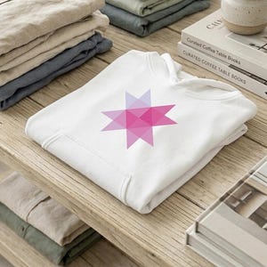 May include: White hoodie with a geometric star design in shades of pink and purple. The sweatshirt is folded on a wooden surface, with stacks of folded clothing in the background. The star design is a prominent feature.