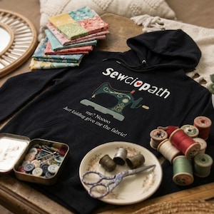 Könnte beinhalten: Ein schwarzer Kapuzenpullover mit dem Text "Sewciopath" und einer Nähmaschinen-Grafik. Das Bild zeigt auch Stoffquadrate, Garnrollen, Knöpfe, Nadeln und Scheren, alles im Zusammenhang mit Nähen und Basteln.