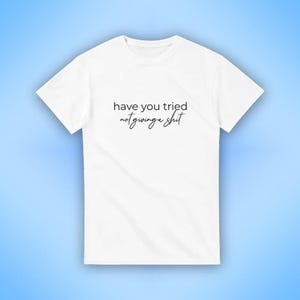 Puede incluir: Camiseta blanca con la frase "have you tried not giving a shit" impresa en escritura negra. La camiseta se muestra sobre un fondo azul claro. La camiseta tiene cuello redondo y mangas cortas.