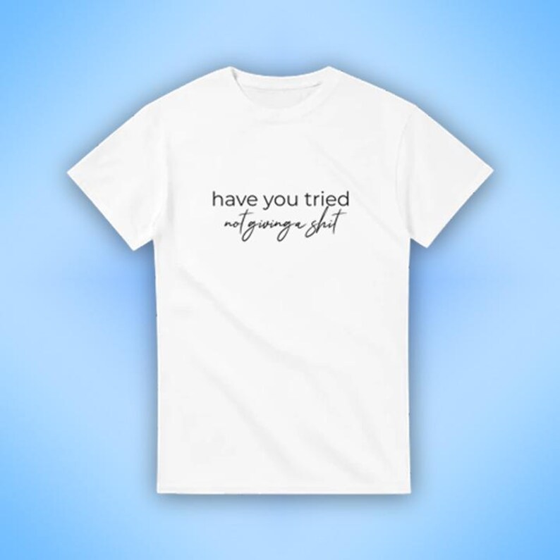 Puede incluir: Camiseta blanca con la frase "have you tried not giving a shit" impresa en escritura negra. La camiseta se muestra sobre un fondo azul claro. La camiseta tiene cuello redondo y mangas cortas.