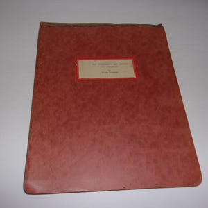 May include: A rectangular, reddish-brown document folder with a textured surface. Centered on the front is a rectangular white label with a thin red border, featuring the printed text: THE HISTORY OF DATES OF AMERICA BY ALICE PERRY.