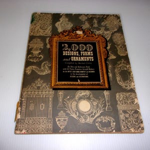 May include: An antique book titled "2,000 Designs, Forms and Ornaments" with a gold-framed title box. The cover is dark gray with ornate illustrations in gold. The book is slightly worn, showing signs of age.