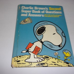 May include: A hardcover book titled Charlie Brown's Second Super Book of Questions and Answers. The blue cover features Snoopy in a space helmet on the moon, holding a red book, with Woodstock nearby and a small space capsule above.