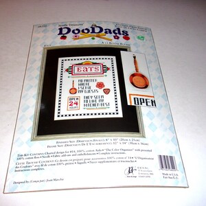 May include: A cross-stitch kit by Needle Treasures, featuring a framed design with the word "EATS" and kitchen-themed motifs. The kit includes a smaller image of a pan and the word "OPEN". The finished size is 8" x 10".