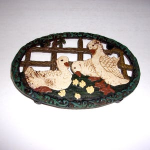 May include: An oval cast iron trivet featuring two cream-colored ducks positioned in front of a brown lattice fence. The scene includes small yellow flowers on a dark green ground, all enclosed within a decorative dark green and black border.