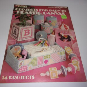 May include: A Leisure Arts craft booklet titled Projects for Baby in Plastic Canvas featuring 14 projects. The cover displays various plastic canvas items including a nursery organizer, baby blocks, a mobile, and a rattle in pastel colors.