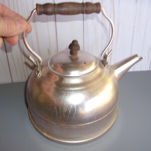 May include: A vintage silver-colored tea kettle with a wooden handle and a spout. The kettle has a round body and a lid with a small wooden knob. The handle is arched and made of metal. The kettle is designed for heating water.