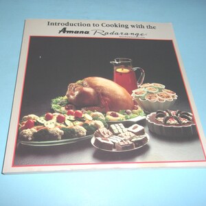 May include: A cookbook titled "Introduction to Cooking with the Amana Radarange Microwave Oven" featuring a roasted turkey, side dishes, and desserts. The cover has a red border and a blue background.
