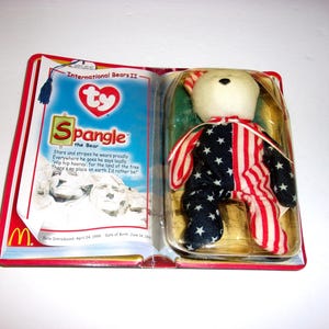 May include: A Ty Spangle the Bear plush toy in a plastic blister pack. The bear has a cream-colored head and a body patterned with red and white stripes and blue fabric with white stars. The packaging features the text International Bears II.