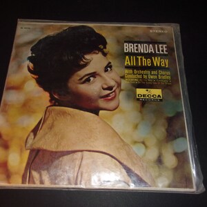 May include: Vintage vinyl record album cover for Brenda Lee's "All The Way." The cover features a smiling woman with dark hair and a light-colored coat. The album title and artist's name are in bold text. The Decca Records logo is visible.