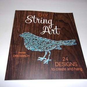 May include: A softcover book titled DIY String Art by Jesse Dresbach with a dark brown wood grain background. The cover features a light blue string art bird graphic and the text 24 DESIGNS to create and hang in orange and white lettering.