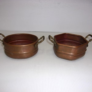 May include: Two copper pots with brass handles. One is round, the other is hexagonal. Both have a reddish-brown interior and a shiny copper exterior. The pots are sitting on a white surface.