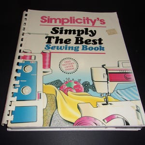May include: A spiral-bound instructional sewing book titled Simplicity's Simply The Best Sewing Book. The cover features illustrations of sewing machines, a pink pincushion, and yellow fabric, with a circular badge stating Free Pattern Offer Inside.