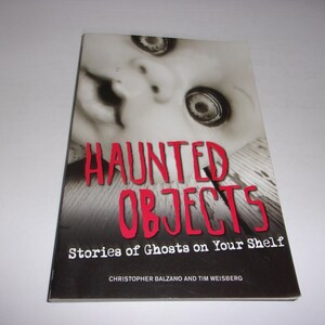 May include: A paperback book titled Haunted Objects: Stories of Ghosts on Your Shelf by Christopher Balzano and Tim Weisberg. The cover features a black and white close-up of a doll face with large eyes and bold red text for the title.