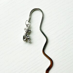 May include: A silver-toned metal bookmark featuring a wavy, serpentine design. Attached to the top loop of the bookmark is a small, detailed charm depicting a seated, humanoid figure with pointed ears and a smooth, reflective metallic finish.