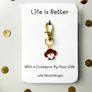 May include: A gold-tone keychain featuring a brown and white Cockapoo charm. The keychain hangs from a white card with the text "Life is Better With a Cockapoo By Your side" and "Julie Davina Designs".