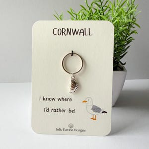 May include: A silver-toned ring with a small seashell charm attached, displayed on a cream-coloured card featuring the text "CORNWALL", "I know where I'd rather be!", and "Julie Davina Designs" alongside a graphic illustration of a seagull.