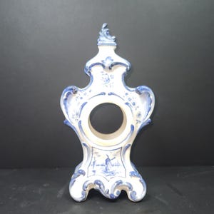 Op de afbeelding: Een witte keramische klokkenstandaard met blauwe en witte bloemmotieven. De standaard heeft een windmolenscène en een ronde opening voor een wijzerplaat.