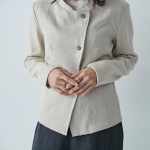 May include: A light beige jacket with a mandarin collar and two dark gray buttons. The jacket has a wrap-style front and long sleeves. The person is wearing silver rings. The jacket is made of a textured fabric.