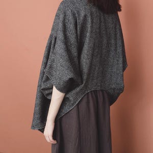 Peut inclure: Un haut de style poncho gris foncé surdimensionné avec un motif en chevrons. Le haut a des manches larges et une silhouette drapée. Le modèle porte également une jupe marron foncé. Le fond est orange uni et discret.