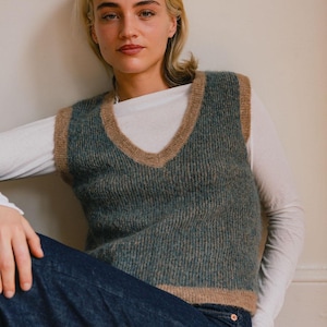 May include: A teal and tan V-neck sweater vest. The vest is worn over a white long-sleeve shirt and paired with dark blue jeans. The model is sitting and leaning against a wall.