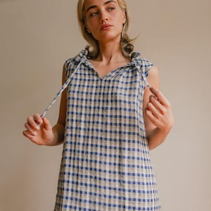 May include: Sleeveless blue and white gingham dress with a V-neckline and tie details at the shoulders. The dress has a relaxed fit and a classic checkered pattern, perfect for casual wear.