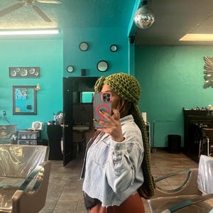 以下が含まれることがあります： 緑色の編み込みのヘッドラップと長いドレッドヘア、薄いブルーのデニムジャケット、茶色のズボンを着用した人がいます。その人は美容院で自撮りしています。