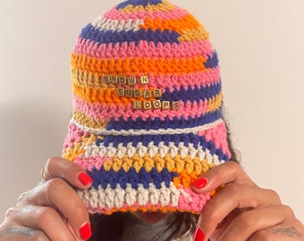May include: A colorful crochet bucket hat with the text "BROWN SUGAR LOOPS" in gold letters. The hat features a vibrant mix of pink, orange, yellow, blue, and white yarn. The hat is handmade and has a unique, textured appearance.