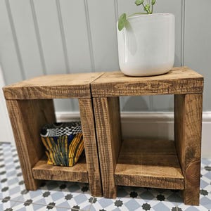 May include: Two wooden shelves with a natural brown finish. The shelves are stacked on top of each other and have a square shape. The top shelf has a white ceramic pot with a green plant. The bottom shelf has a colourful fabric box.
