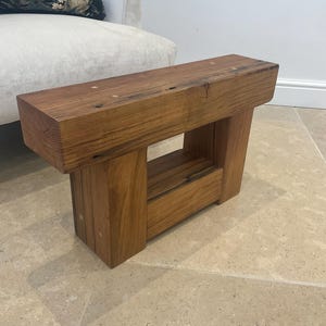 May include: A solid wooden bench with a natural finish. The bench features a thick, rectangular top supported by two vertical legs, with a rectangular space in the middle. The wood grain is visible, and the bench appears to be made from reclaimed timber.