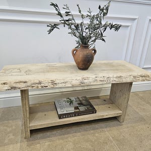 May include: A light-toned wooden coffee table with a lower shelf. A small, brown vase with olive branches sits on top. A book with the word "CREATE" is on the shelf. The table has a natural, unfinished appearance.