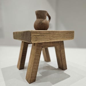 May include: A small wooden stool with four legs and a square top. A small brown clay jug sits on top of the stool.
