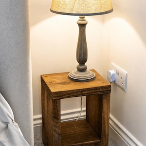 May include: A wooden bedside table with a lamp. The table is a square, open-shelf design with a dark brown finish. A gray lamp with a beige shade sits on top. The lamp has a gray base. The table is near a wall with an electrical socket.
