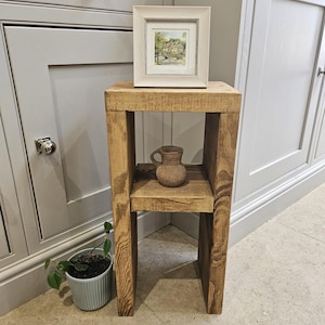 May include: A wooden side table with a natural finish, featuring a square design and a lower shelf. A framed picture of a landscape scene sits on top. A small ceramic jug is on the shelf. A potted plant is next to the table.