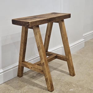 May include: A wooden stool with a rectangular seat and A-frame legs. The stool is constructed from light brown wood, showcasing the natural grain and nail fixings. The design is simple and robust, suitable for various uses.