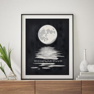 May include: A framed wall art print featuring a full moon centered against a dark, textured black background. Below the moon, a white reflection shimmers on a surface, accompanied by the white cursive text, your custom text goes here.