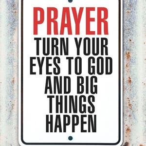 May include: A rectangular white sign with a black border and the word "PRAYER" in red at the top. Below, the sign reads "TURN YOUR EYES TO GOD AND BIG THINGS HAPPEN" in black text. The sign is mounted on a weathered, corrugated metal surface.