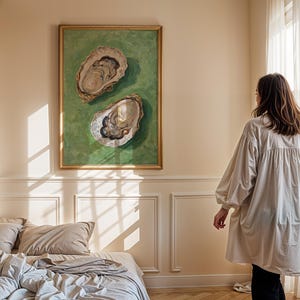 Op de afbeelding: Een ingelijst schilderij van twee oesters op een groene achtergrond hangt aan een muur in een slaapkamer. Het schilderij is in een gouden lijst. Een vrouw in een wit shirt staat in de kamer en kijkt naar het schilderij.