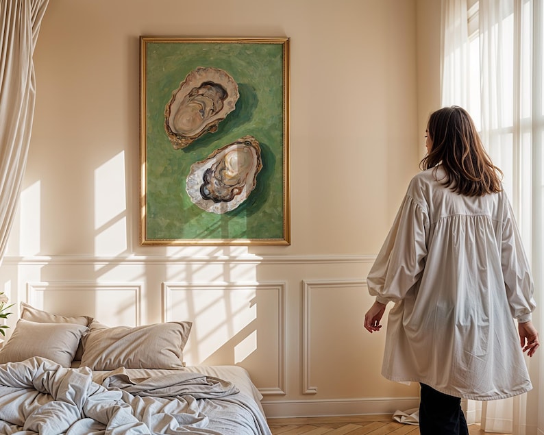 Op de afbeelding: Een ingelijst schilderij van twee oesters op een groene achtergrond hangt aan een muur in een slaapkamer. Het schilderij is in een gouden lijst. Een vrouw in een wit shirt staat in de kamer en kijkt naar het schilderij.