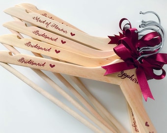 Puede incluir: Perchas de madera clara con texto grabado y detalles de corazón. Las perchas presentan las palabras "Dama de honor" y "Dama de honor" en una fuente cursiva. Una cinta y un lazo de color burdeos adornan las perchas.