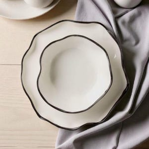 Può includere: Due piatti in ceramica bianca con bordo ondulato e rifinitura grigio scuro. I piatti sono impilati, con un panno grigio sullo sfondo. I piatti fanno parte di un set di stoviglie.