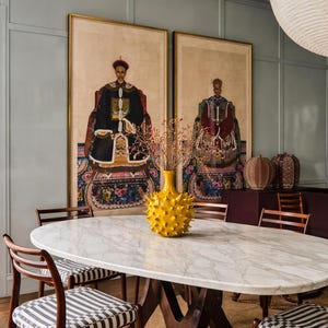 Könnte beinhalten: Ein Esszimmer mit einem ovalen Marmortisch und gestreiften Stühlen. Zwei gerahmte Porträts hängen an der Wand, und eine gelbe, stachelige Vase steht auf dem Tisch. Der Raum hat eine neutrale Farbpalette. Der Tisch ist etwa 200 cm lang.