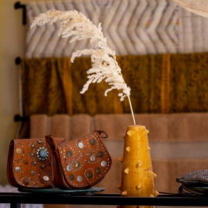 May include: Two brown leather saddlebags with metal studs and turquoise accents sit on a glass plate. A yellow vase with spike-like protrusions holds a dried plant stem. The background features textured fabric.