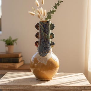 Puede incluir: Un jarrón de cerámica hecho a mano con un diseño único. El jarrón presenta una base bulbosa en un tono amarillo cálido, que transiciona a un cuello azul y verde moteado. Elementos decorativos adornan el cuello. El jarrón contiene flores secas y vegetación, colocado sobre una mesa de madera.