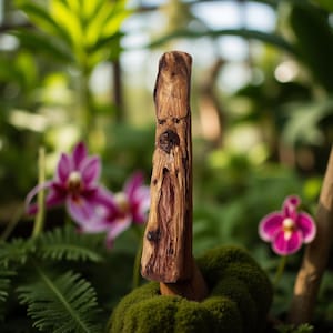 Puede incluir: Una escultura de madera tallada con un acabado natural y texturizado. La escultura marrón, con un tallado detallado en forma de rostro, se alza verticalmente. Se encuentra frente a un fondo borroso de follaje verde y orquídeas rosas.