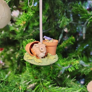 May include: A brown rabbit ornament peeking out of a broken flower pot with a blue bird perched on the rim. The ornament is hanging from a white ribbon on a green Christmas tree.