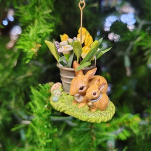 May include: A ceramic ornament featuring two brown bunnies sitting on a green base with a pot of yellow and white flowers. The ornament is hanging from a silver string.