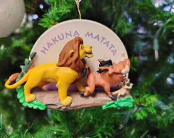 May include: A Christmas ornament featuring Simba and Pumbaa from Disney's The Lion King. The ornament is a round, white disc with the words "Hakuna Matata" written in purple. Simba and Pumbaa are depicted in a 3D design, standing on a brown log with green leaves.