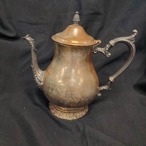 May include: Antique silver-plated teapot with a copper-toned lid and ornate handle and spout. The teapot has a bulbous body and sits on a decorative base. The lid is topped with a small, decorative finial. The teapot is likely from the early 20th century.