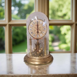 May include: An antique brass clock with a glass dome. The clock face features Roman numerals and ornate detailing. The pendulum and weights are visible inside the glass. The clock sits on a round brass base. The background shows a window with a view of greenery.